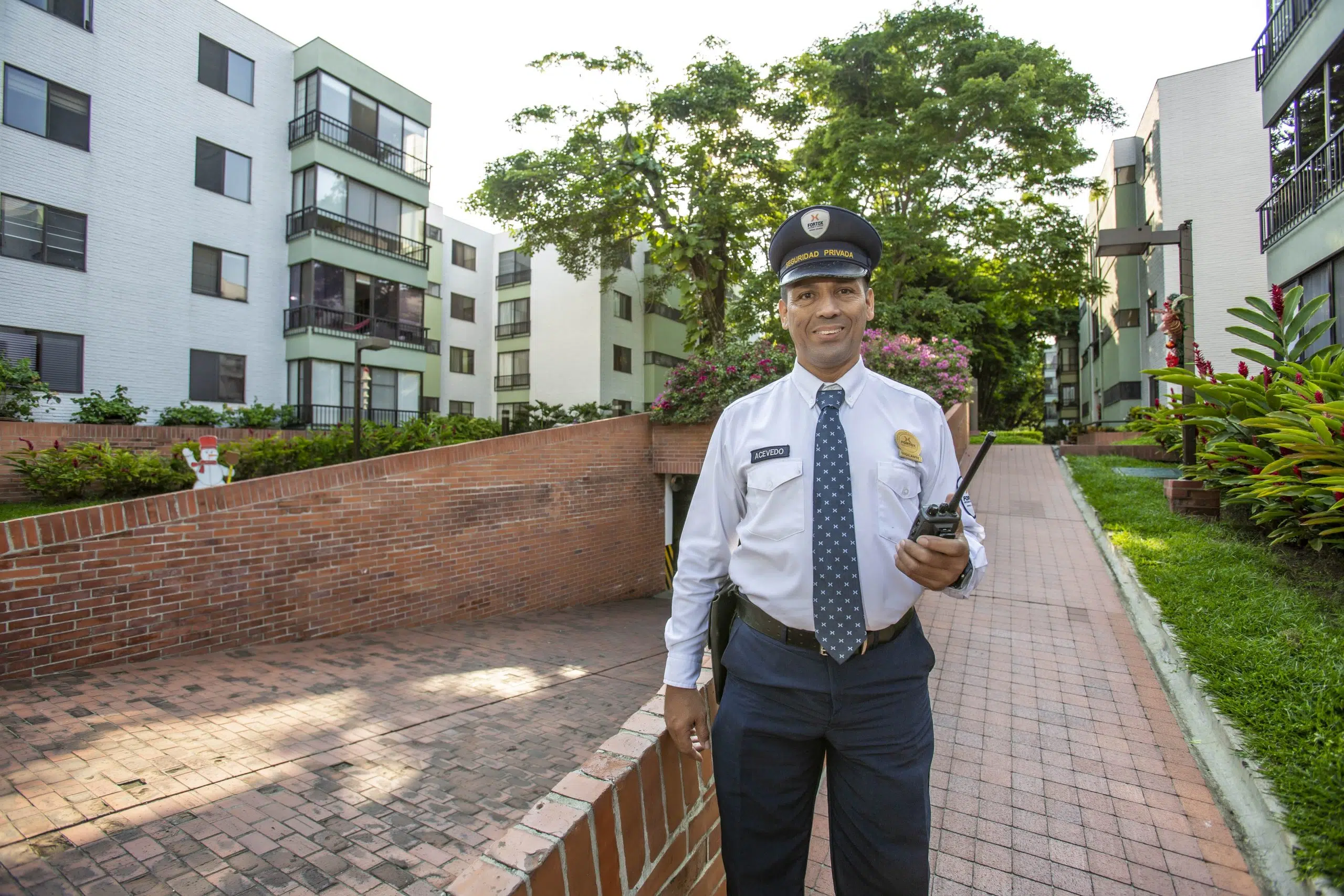 En la imagen se ve un guardia de seguridad privada uniformado de Fortox, sosteniendo una radio de comunicación y una carpeta. El guardia está de pie sobre un sendero de ladrillo, frente a varios edificios residenciales, lo que resalta el servicio de vigilancia y protección empresarial ofrecido por la empresa. - Fortox Security