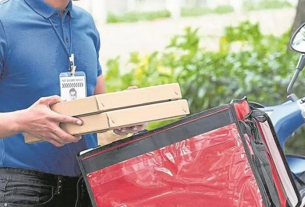 En la imagen, una persona viste una camiseta azul y sostiene dos cajas de pizza mientras se dispone a colocarlas en un bolso térmico rojo para entregas. No aparecen elementos relacionados con seguridad privada, vigilancia u operación de Fortox Security Group en la escena. - Fortox Security