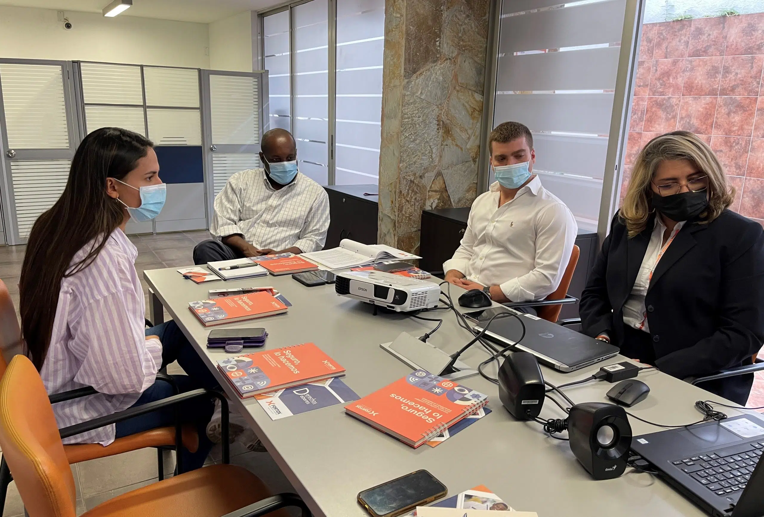 Cuatro personas con tapabocas están sentadas alrededor de una mesa en una oficina moderna, revisando documentos y usando portátiles durante una reunión. Sobre la mesa también hay un proyector. La imagen muestra un ambiente profesional donde el equipo de Fortox Security Group planea estrategias de seguridad privada y vigilancia empresarial. - Fortox Security