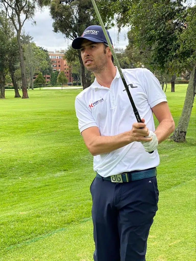 Una persona vestida con polo blanco y pantalón azul oscuro sostiene un palo de golf a medio swing en un campo de golf verde, rodeado de árboles y edificios al fondo. No se observa personal ni vehículos de Fortox Security Group ni elementos relacionados con servicios de seguridad privada o vigilancia en la imagen. - Fortox Security