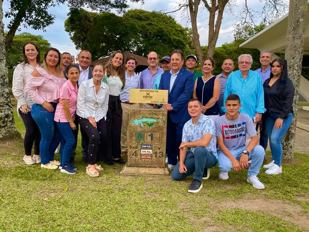 En la imagen se observa a un grupo de unas veinte personas reunidas al aire libre alrededor de una señal de piedra que indica "Tee 1, Par 4, 378 mts, Hoyo 13" en un campo de golf. Al fondo se pueden ver árboles y un edificio. No hay vehículos visibles ni personal uniformado específicamente asociado a seguridad privada o vigilancia de Fortox en la escena. - Fortox Security