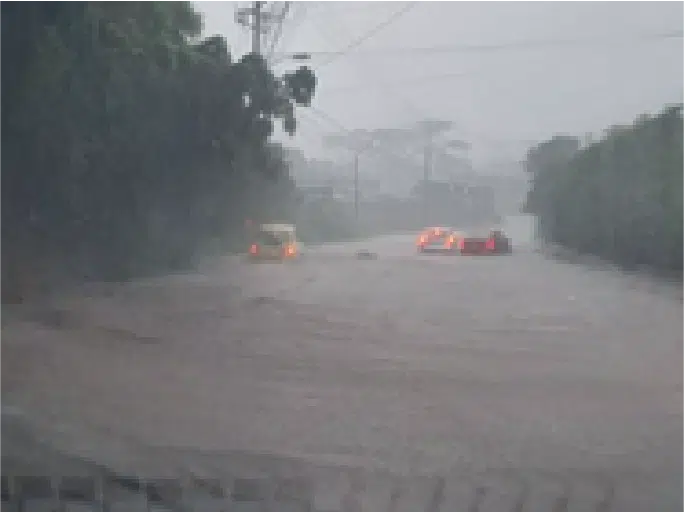 En la imagen se observa una calle inundada debido a lluvias intensas, donde dos vehículos están parcialmente sumergidos en el agua. La visibilidad es limitada por las condiciones climáticas y se pueden ver cables de energía eléctrica colgando encima de la vía. No hay presencia de personal de seguridad privada, vigilancia ni elementos identificables relacionados con Fortox Security Group. - Fortox Security
