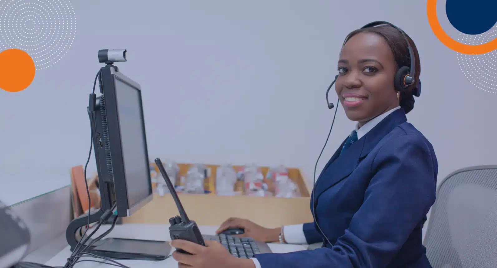 En la imagen se observa a una mujer vestida con un traje azul marino, sentada en un escritorio frente a una computadora. Lleva puesto un auricular y sostiene una radio de comunicación (walkie-talkie) mientras sonríe a la cámara. La escena transmite profesionalismo y está relacionada con servicios de seguridad privada, vigilancia y atención de emergencias, representando posiblemente el trabajo del personal de Fortox Security Group en Colombia. - Fortox Security