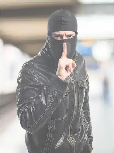 En la imagen se observa a una persona con pasamontañas negras y chaqueta de cuero, llevando un dedo a los labios en señal de silencio. El fondo es un entorno interior desenfocado. La escena puede estar relacionada con acciones que requieren vigilancia o situaciones donde los servicios de seguridad privada como los ofrecidos por Fortox serán necesarios para prevenir robos o intrusiones. - Fortox Security