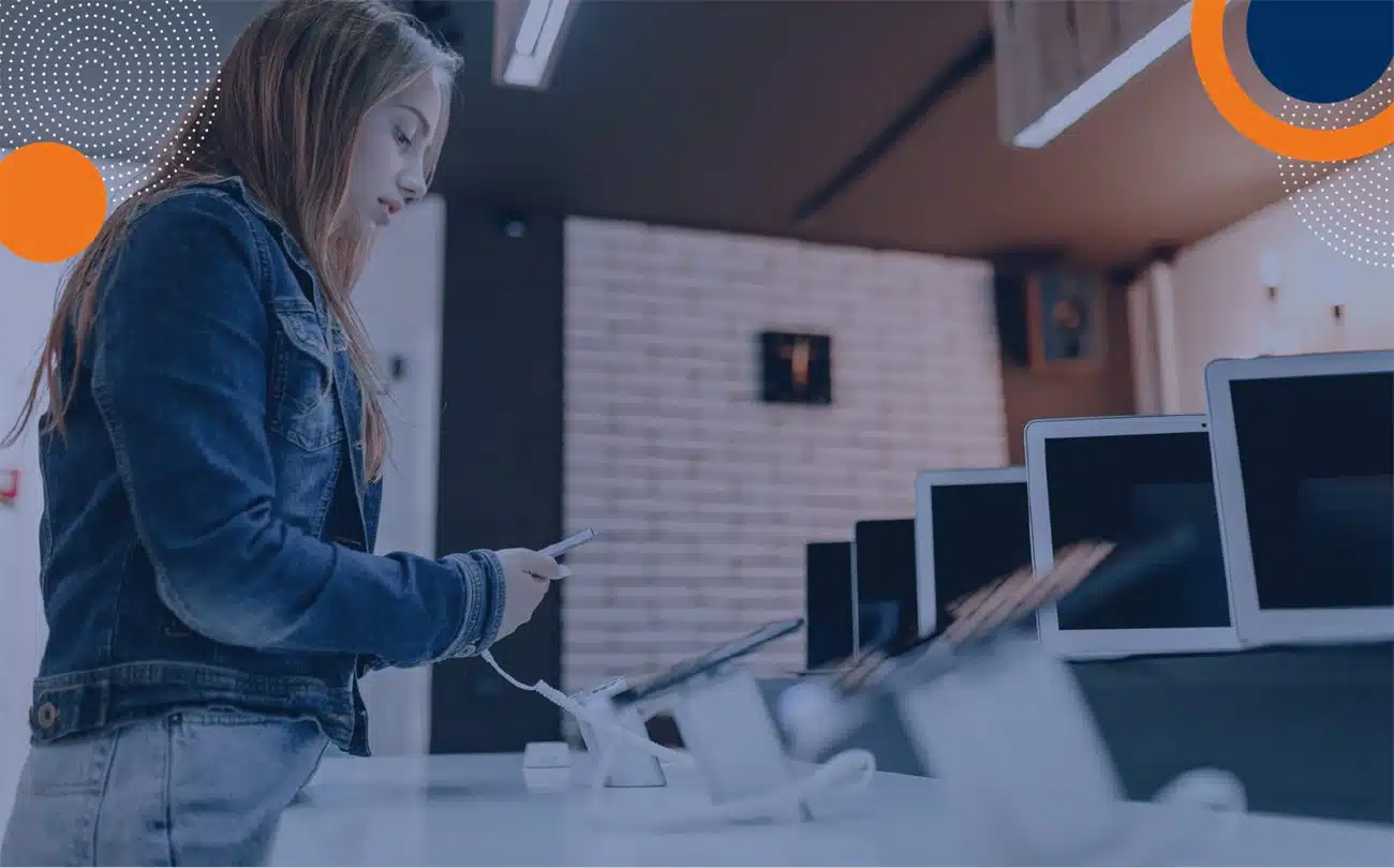 Una persona con chaqueta de mezclilla revisa un teléfono inteligente en una mesa de exhibición dentro de una tienda de dispositivos electrónicos; cerca se observan varias tabletas sobre soportes. No se aprecian elementos relacionados con seguridad privada, vigilancia o Fortox en la imagen. - Fortox Security