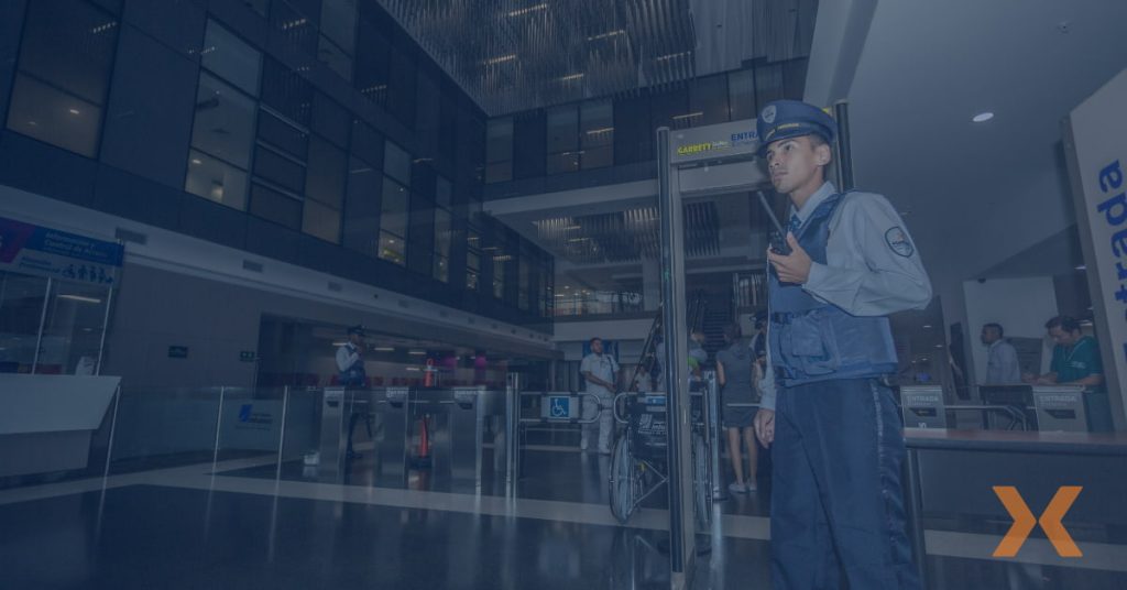 Guardia de seguridad privada de Fortox vigila la entrada de un edificio empresarial moderno junto a detectores metálicos. - Fortox Security
